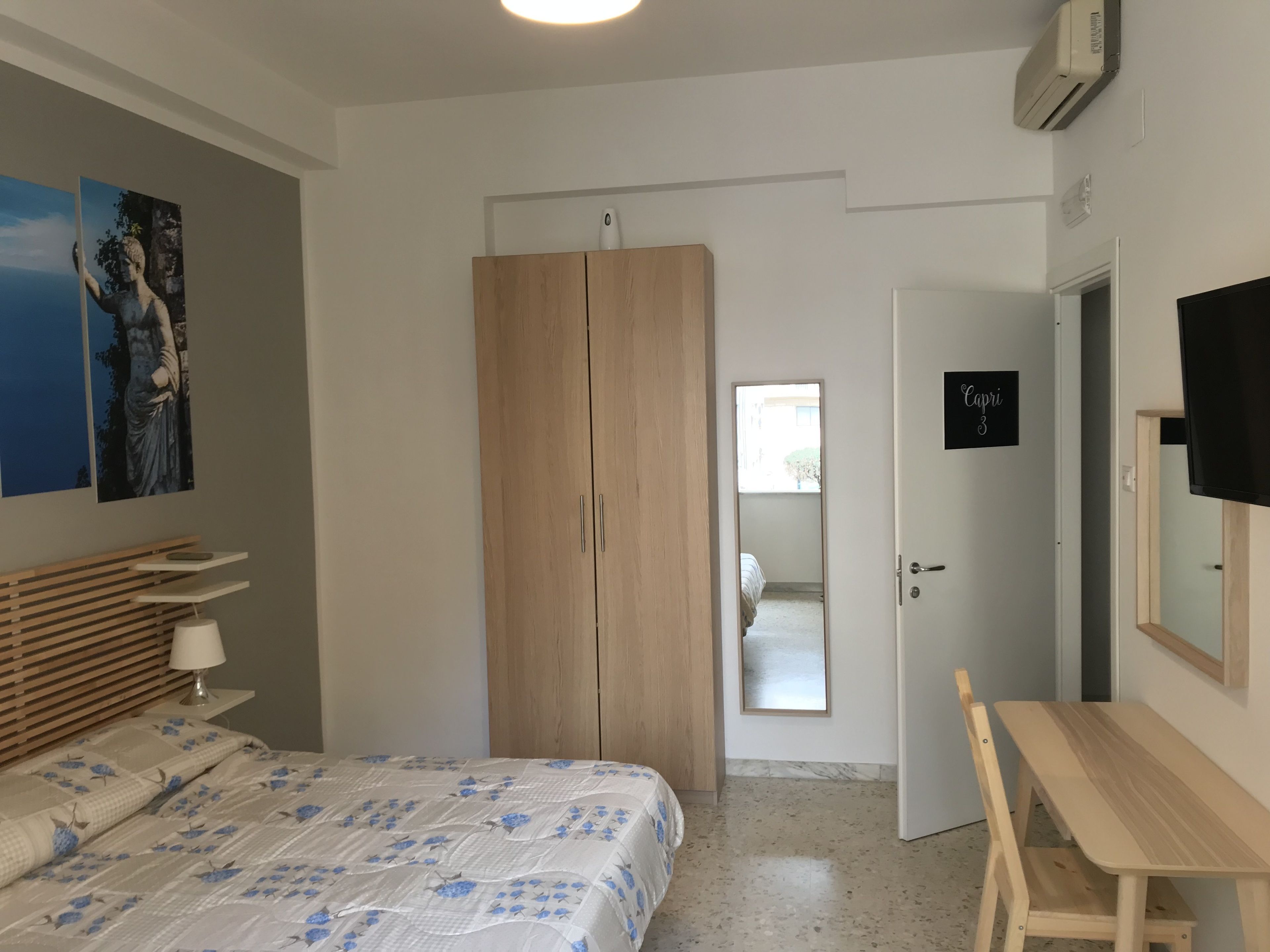 A bedroom with a bed, a wooden wardrobe, a desk, a mirror, and a door labeled "Capri 3".