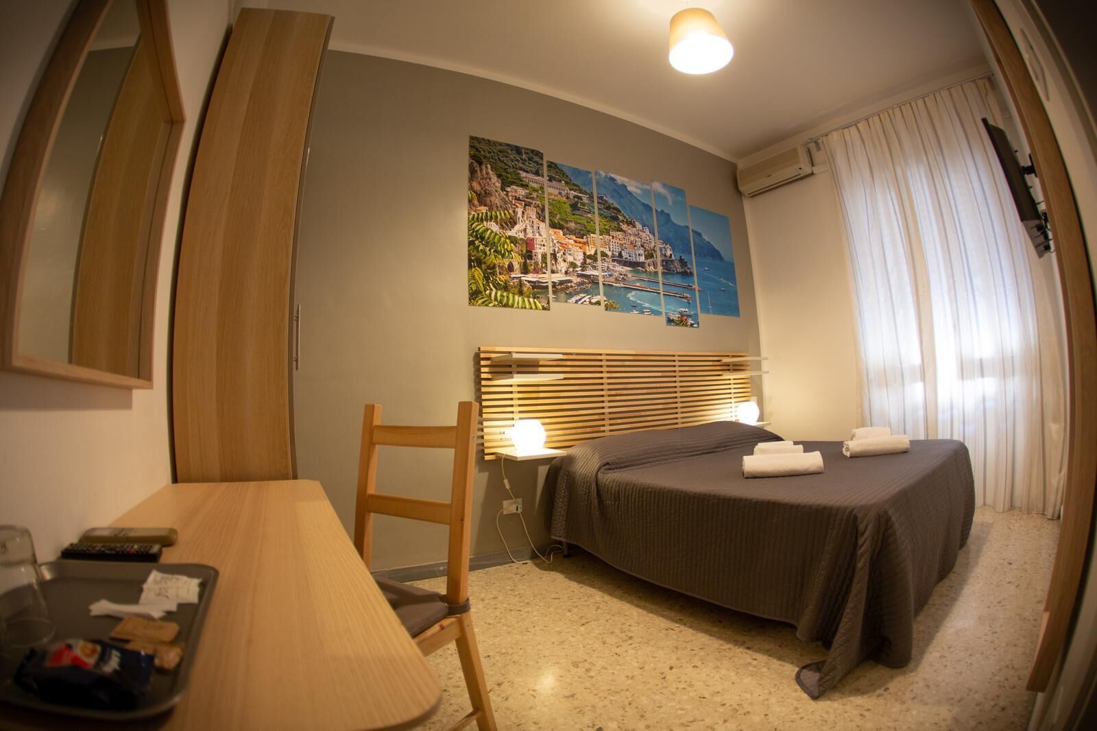 A hotel room with a bed, a desk, a chair, a painting of a coastal town, and a window with curtains.