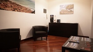 Een kamer met een houten vloer, een zwarte fauteuil, een klein tafeltje met tijdschriften en een koffieapparaat op een dressoir.