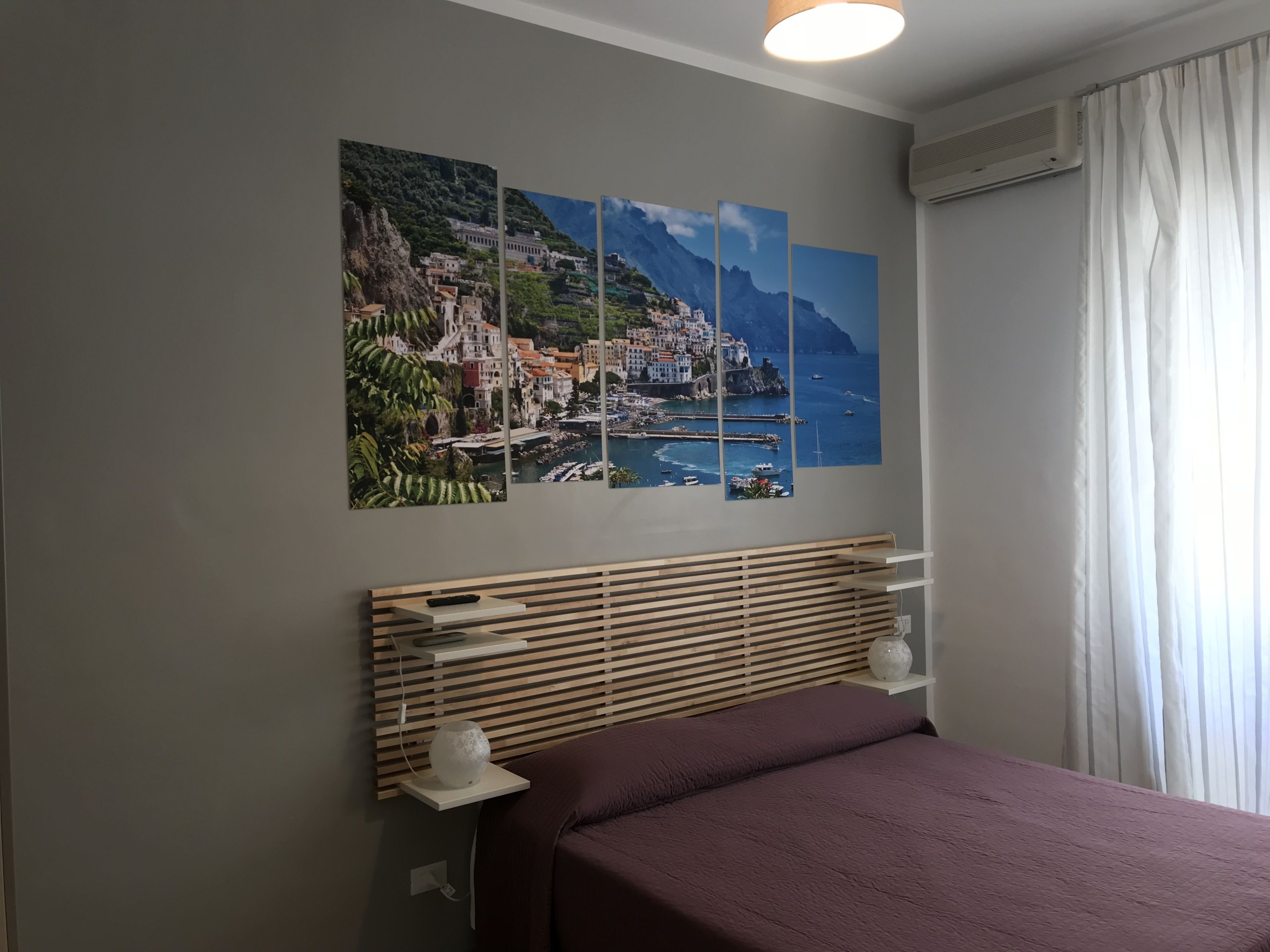 A bedroom with a bed, a wooden headboard, a wall-mounted shelf with a vase, and a large coastal landscape painting.
