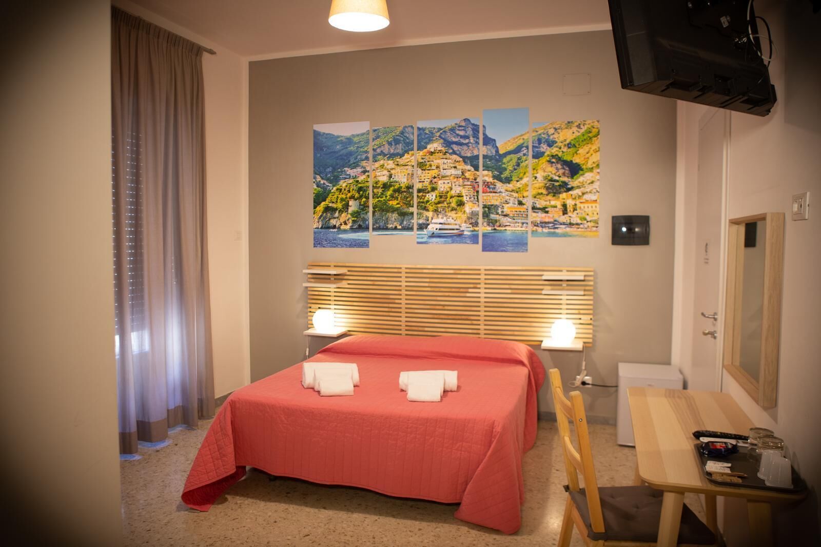 Una camera d'albergo con un letto, una scrivania, una sedia, una TV e un dipinto di un paesaggio costiero.