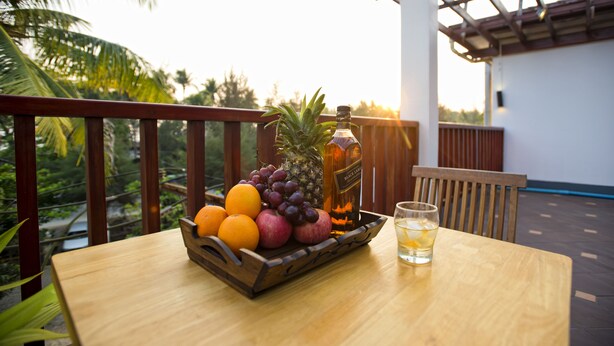 A wooden table with a fruit basket containing oranges, grapes, and a pineapple, a bottle of Black & Decker, and a glass of water on a balcony with a wooden railing and a view of palm trees.