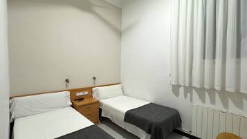 Una habitación de hotel pequeña con dos camas individuales, una mesita de noche de madera, una pequeña mesa y un radiador.