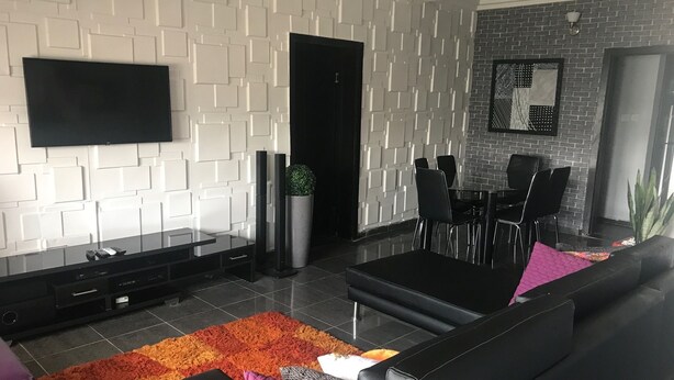 A modern living room with a flat-screen TV, black leather sofa, and a dining area with black chairs and a table. - Grand Condo (Lekki)