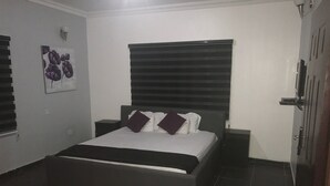 A bedroom with a bed, a nightstand, a wall-mounted light, and a door. - Grand Condo (Lekki)