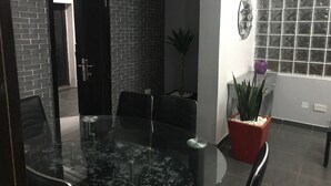 A modern dining area with a glass table surrounded by chairs, a potted plant, and a window with a decorative grille. - Grand Condo (Lekki)