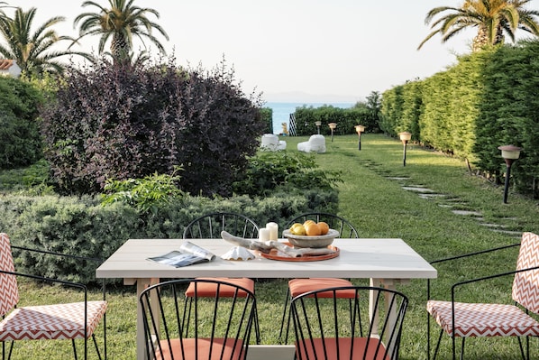 An outdoor dining area with a table and chairs, surrounded by greenery and palm trees, overlooking a body of water.