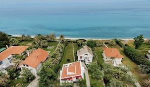 A coastal residential area with houses featuring red-tiled roofs, surrounded by lush greenery and palm trees, with a beach and ocean in the background.