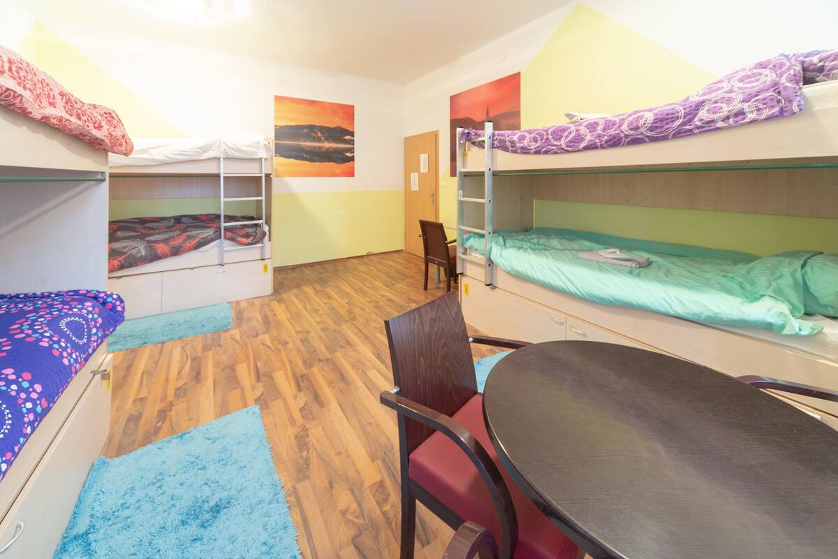 A dormitory room with bunk beds, a desk, and a chair. There are posters on the walls and a door in the background.