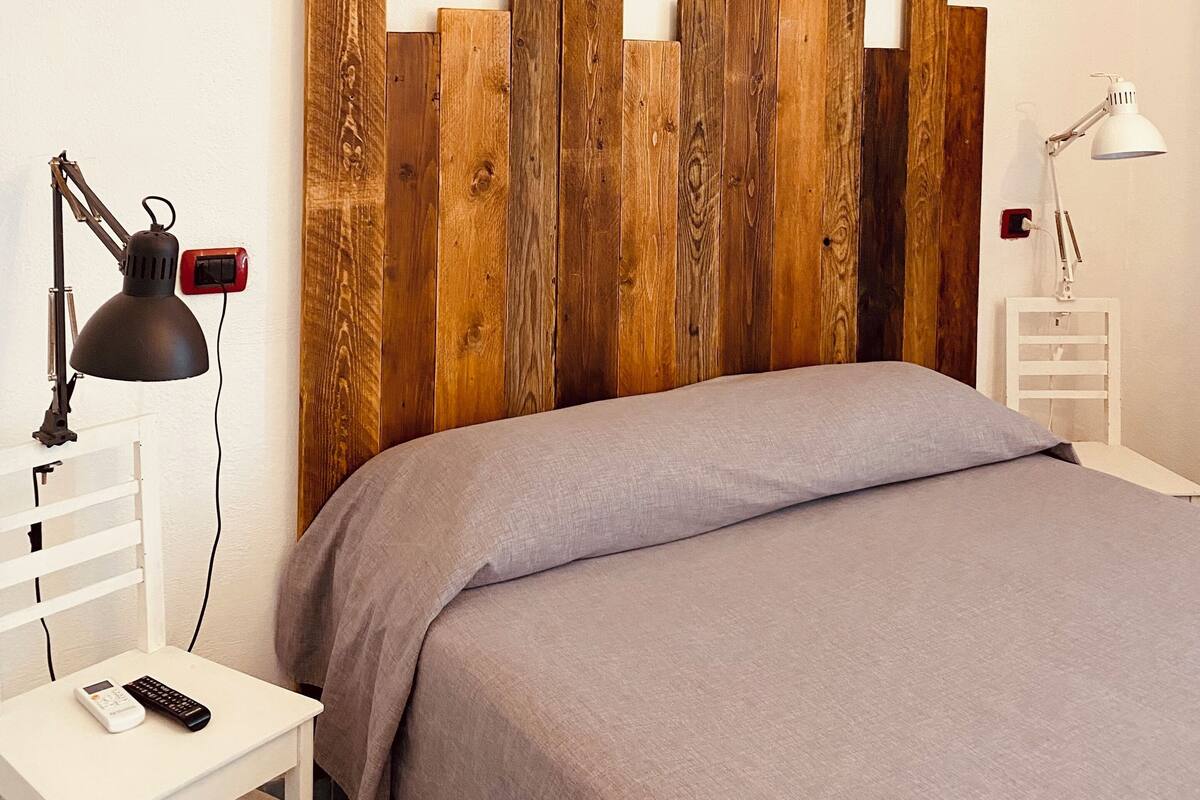 A bedroom with a wooden headboard, a bed with a grey bedspread, a bedside table with a lamp, and a power outlet.