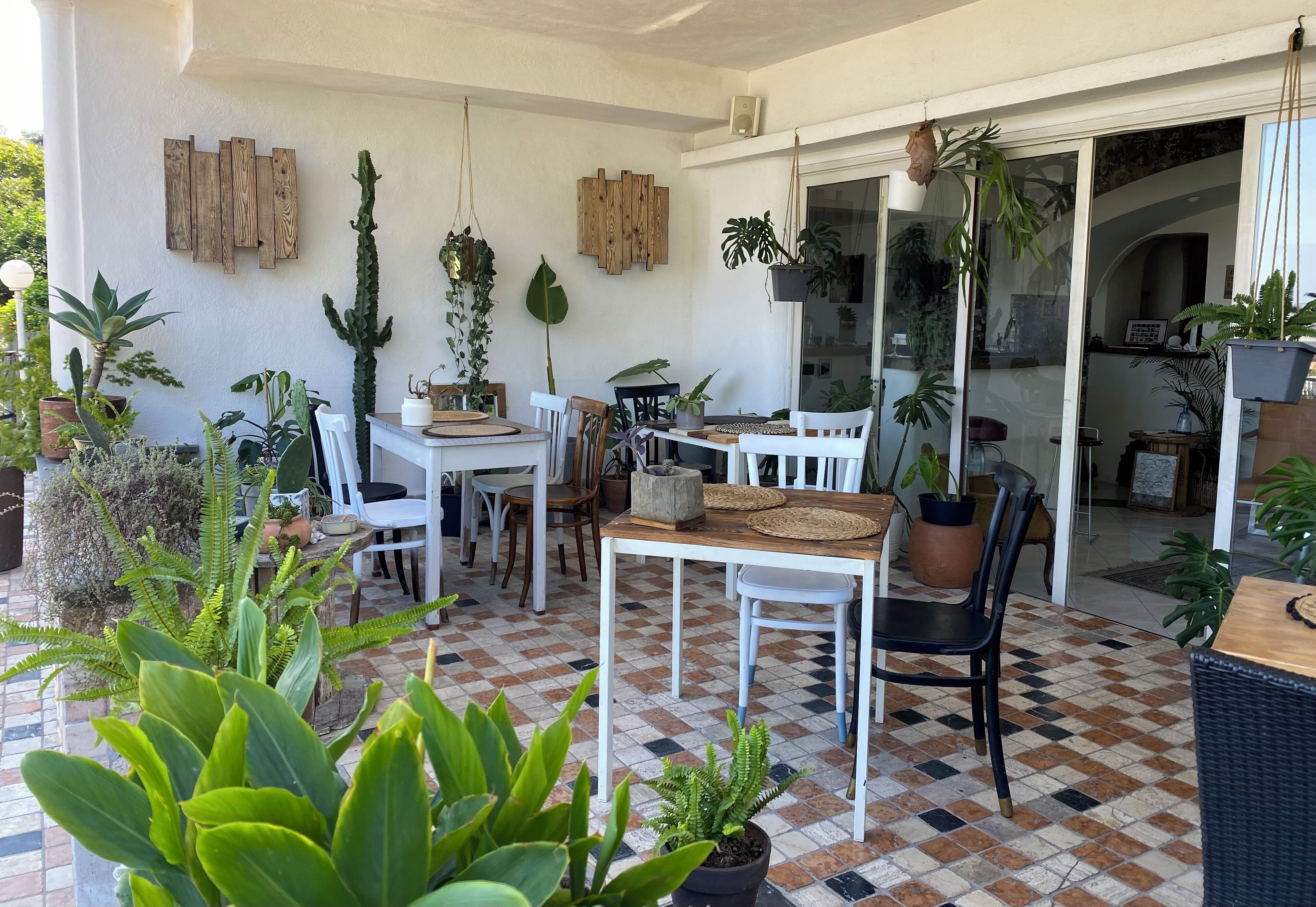 A covered patio area with a checkered floor, white chairs, a wooden table, and various potted plants.