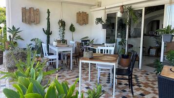 Un patio couvert avec un sol à carreaux, des chaises blanches, une table en bois et diverses plantes en pot.