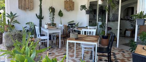 A covered patio area with a checkered floor, white chairs, a wooden table, and various potted plants.