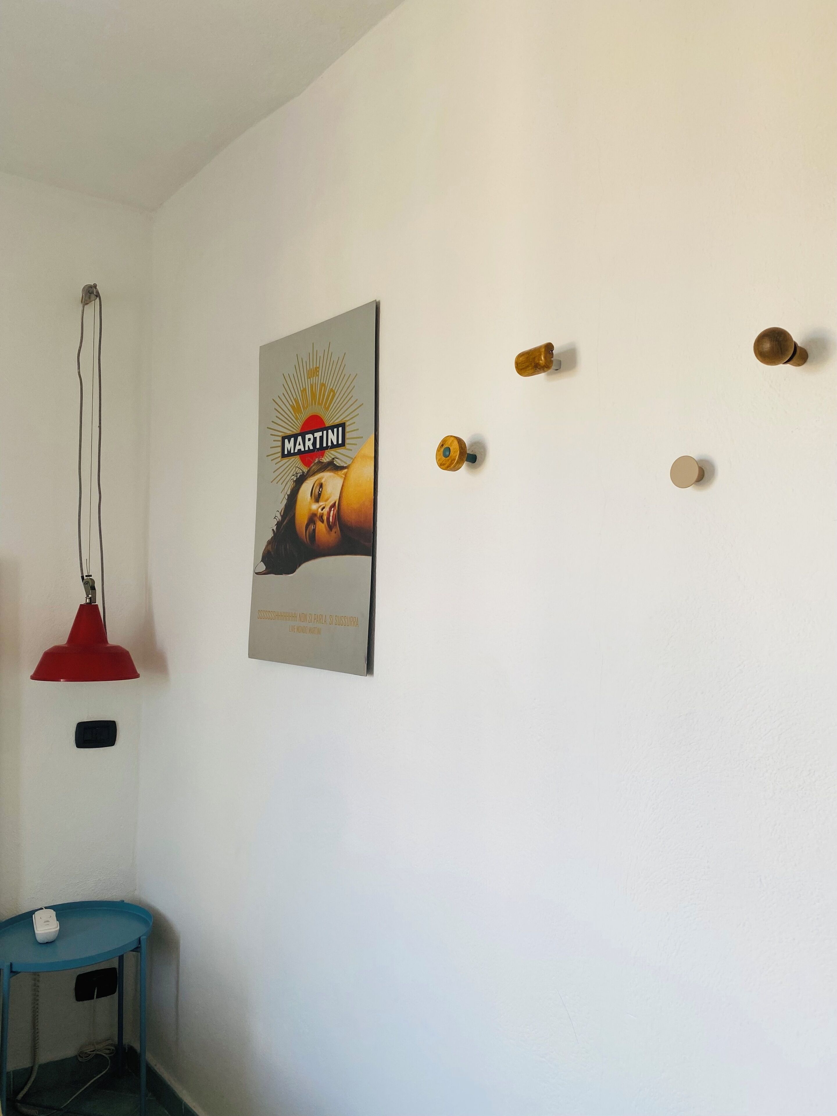 Una stanza con un parete bianca, una lampada a sospensione rossa, un tavolo blu con un telefono bianco e un poster pubblicitario di Martini.