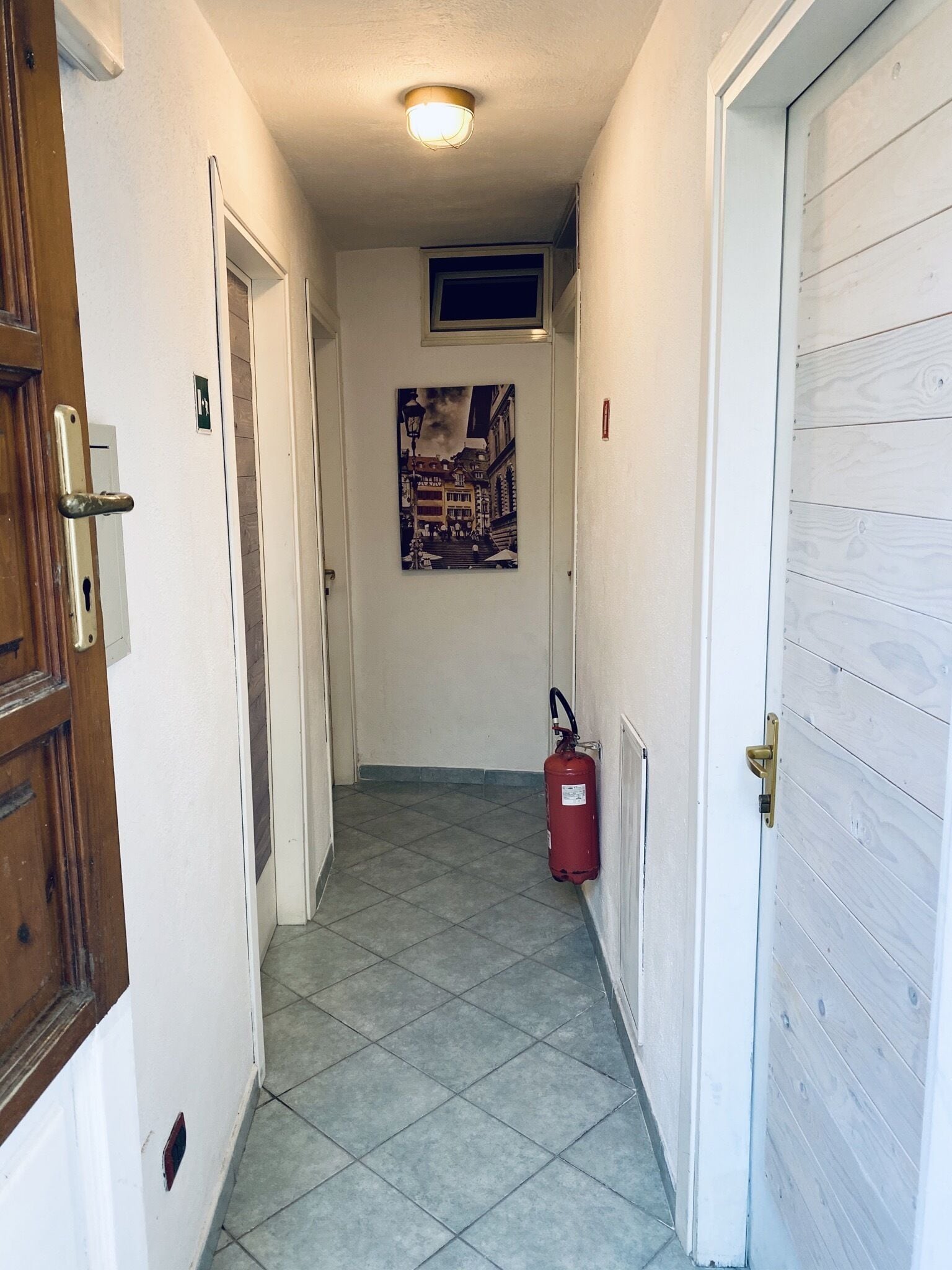 Un pasillo estrecho con un extintor montado en la pared, una puerta con manija y una lámpara en el techo.