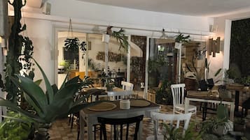 Una stanza con pavimento a scacchi, pareti bianche e diverse piante in vaso. Sono presenti diversi tavoli e sedie disposti per mangiare o sedersi.