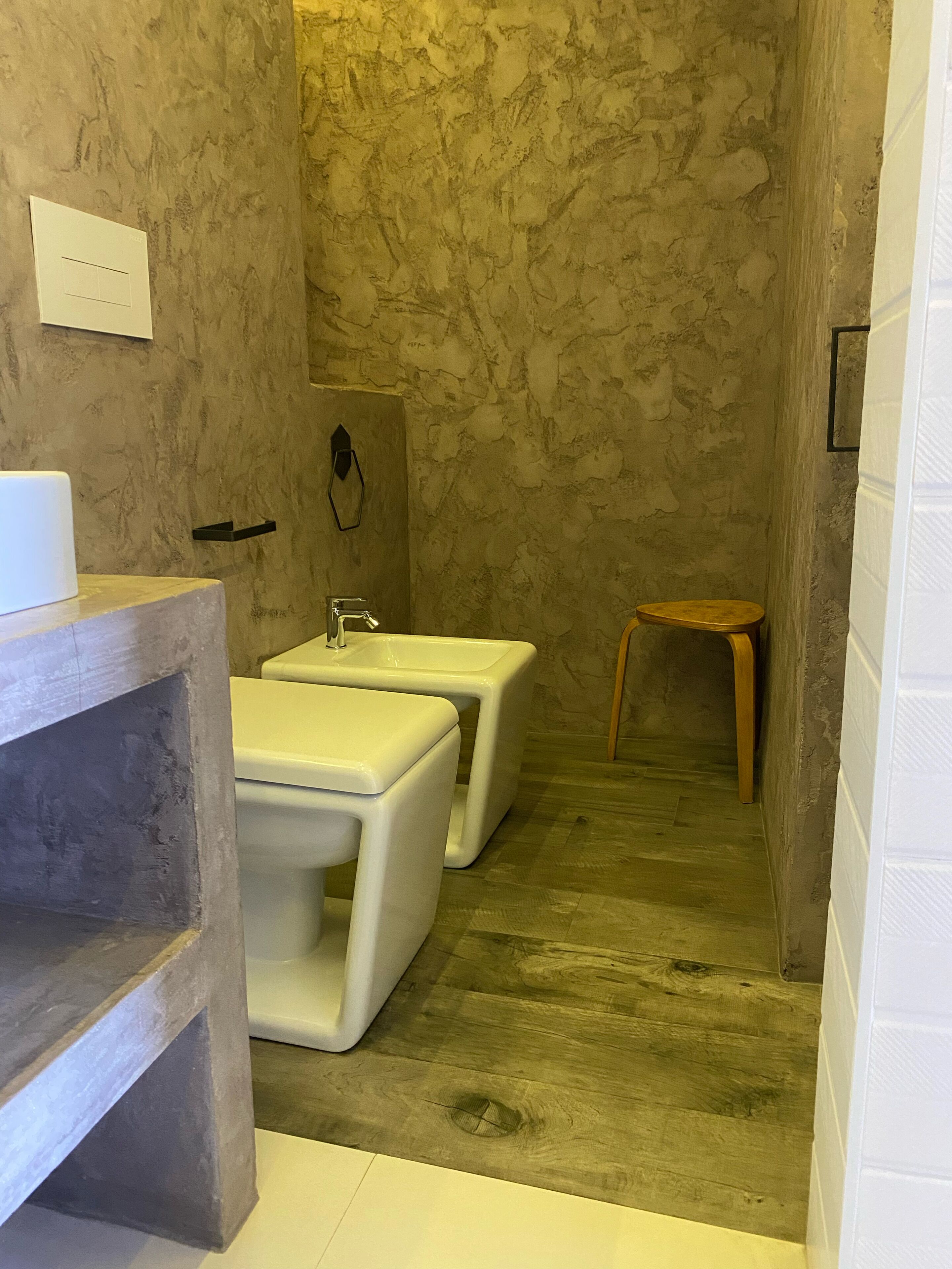 Un baño estrecho con pared de mármol, dos lavamanos blancos, un taburete de madera y un estante blanco.