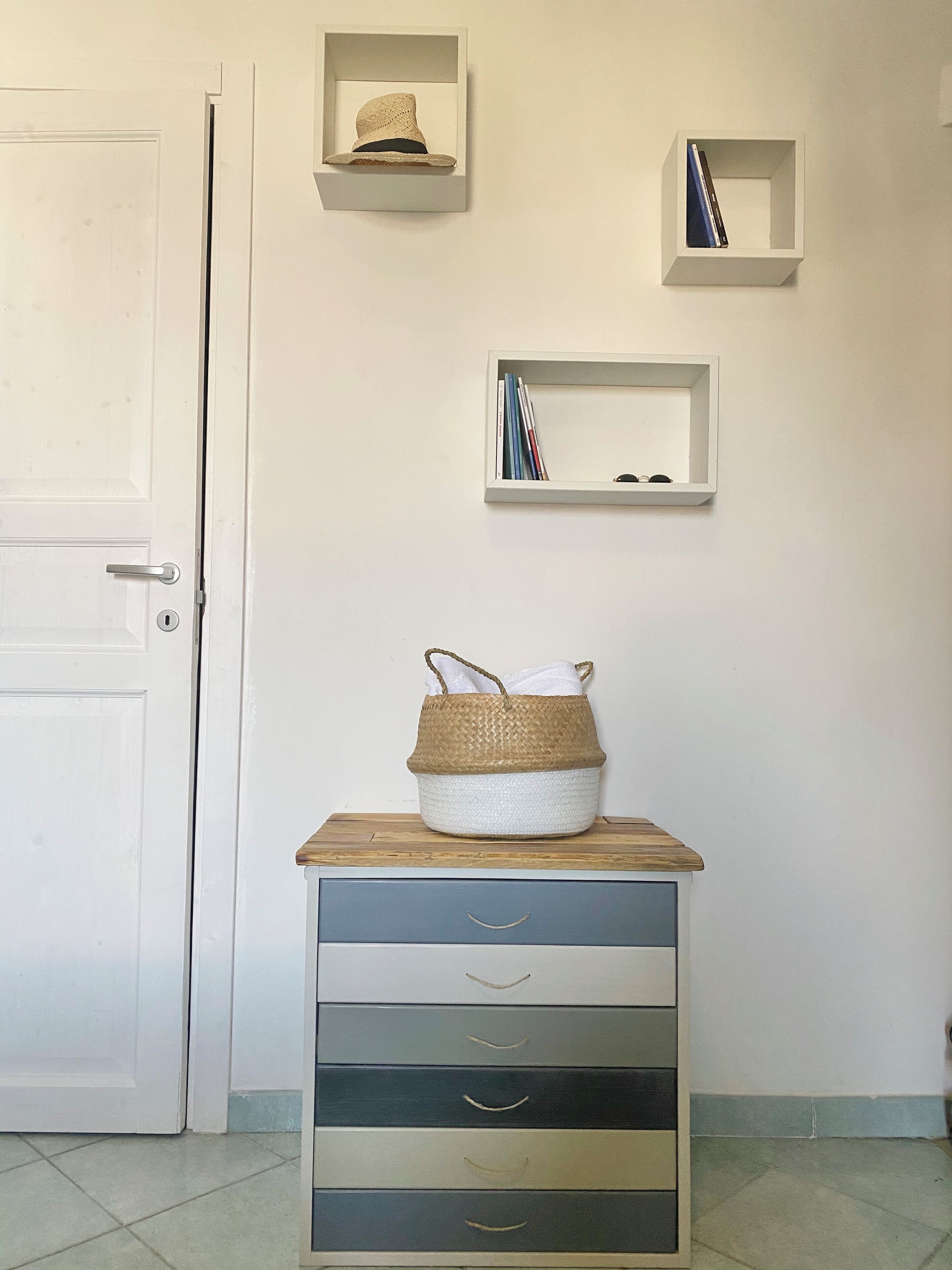 A room with a white door, a wooden cabinet with drawers, and three white shelves on the wall.
