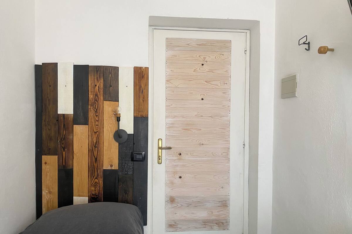 A bedroom with a bed, a door with a wooden panel design, a bedside table, and a wall-mounted light fixture.