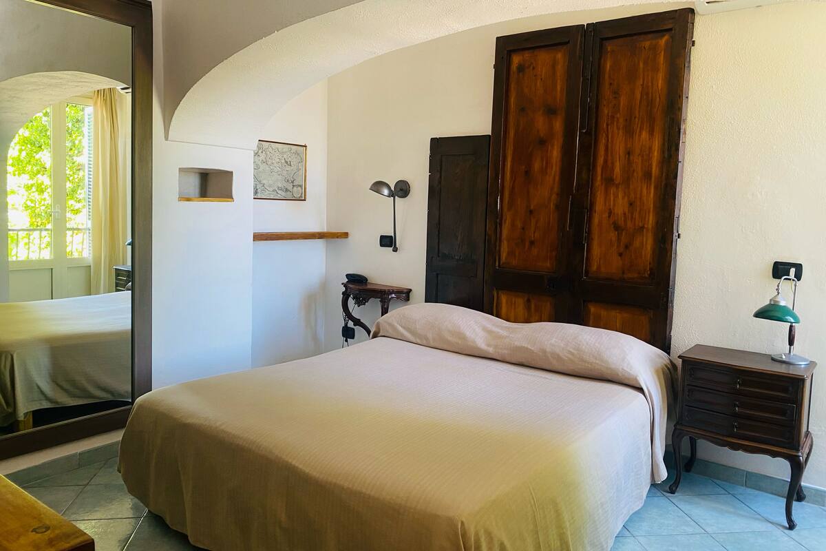 A bedroom with a large bed, a wooden wardrobe, a desk with a lamp, and a mirror on the wall.