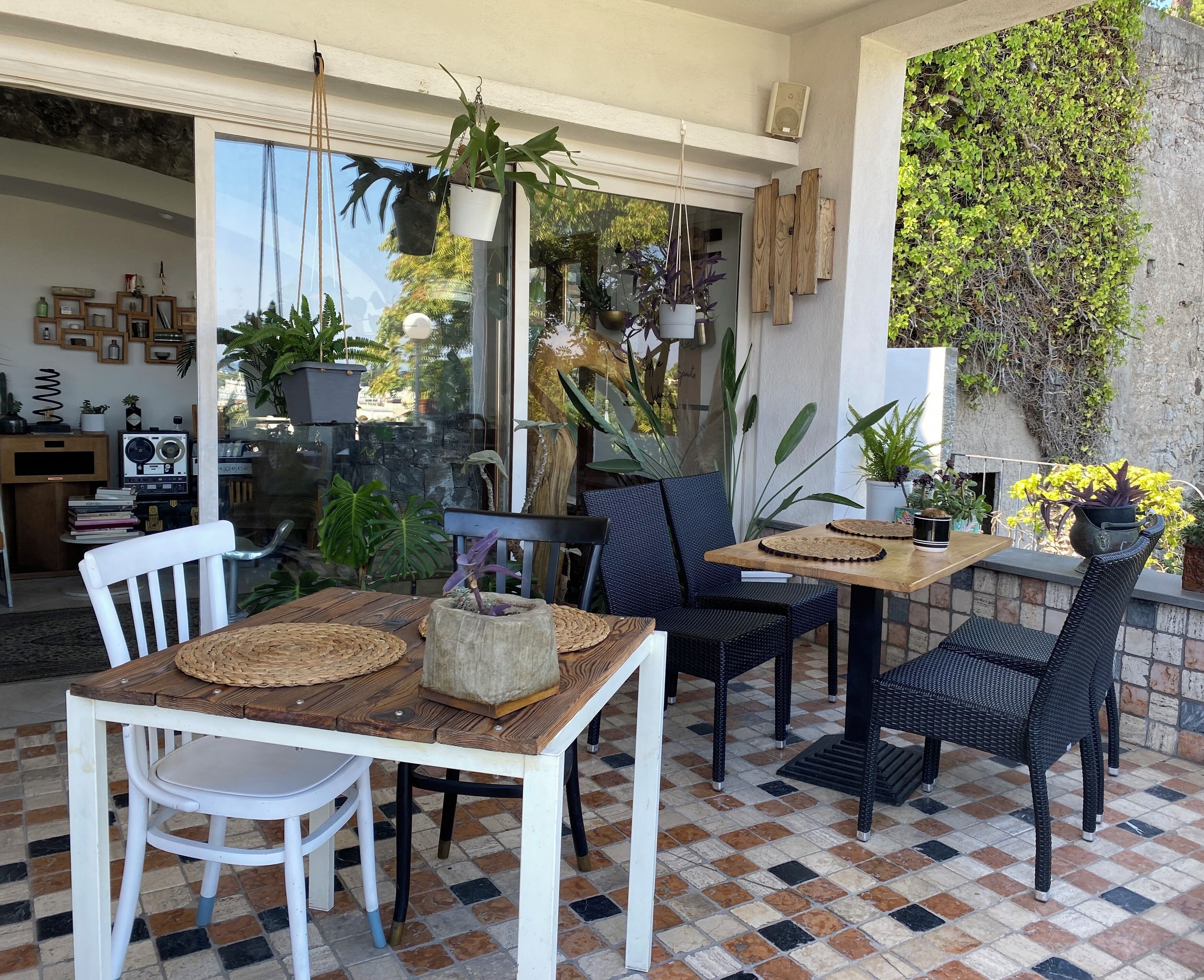 A patio area with a wooden table, two wicker chairs, and a white chair. There are potted plants and a wall with climbing plants.