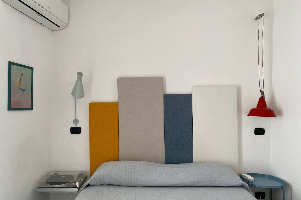 A bed with a grey sheet, a bedside table with a lamp, a phone, and a small shelf with books and a radio.