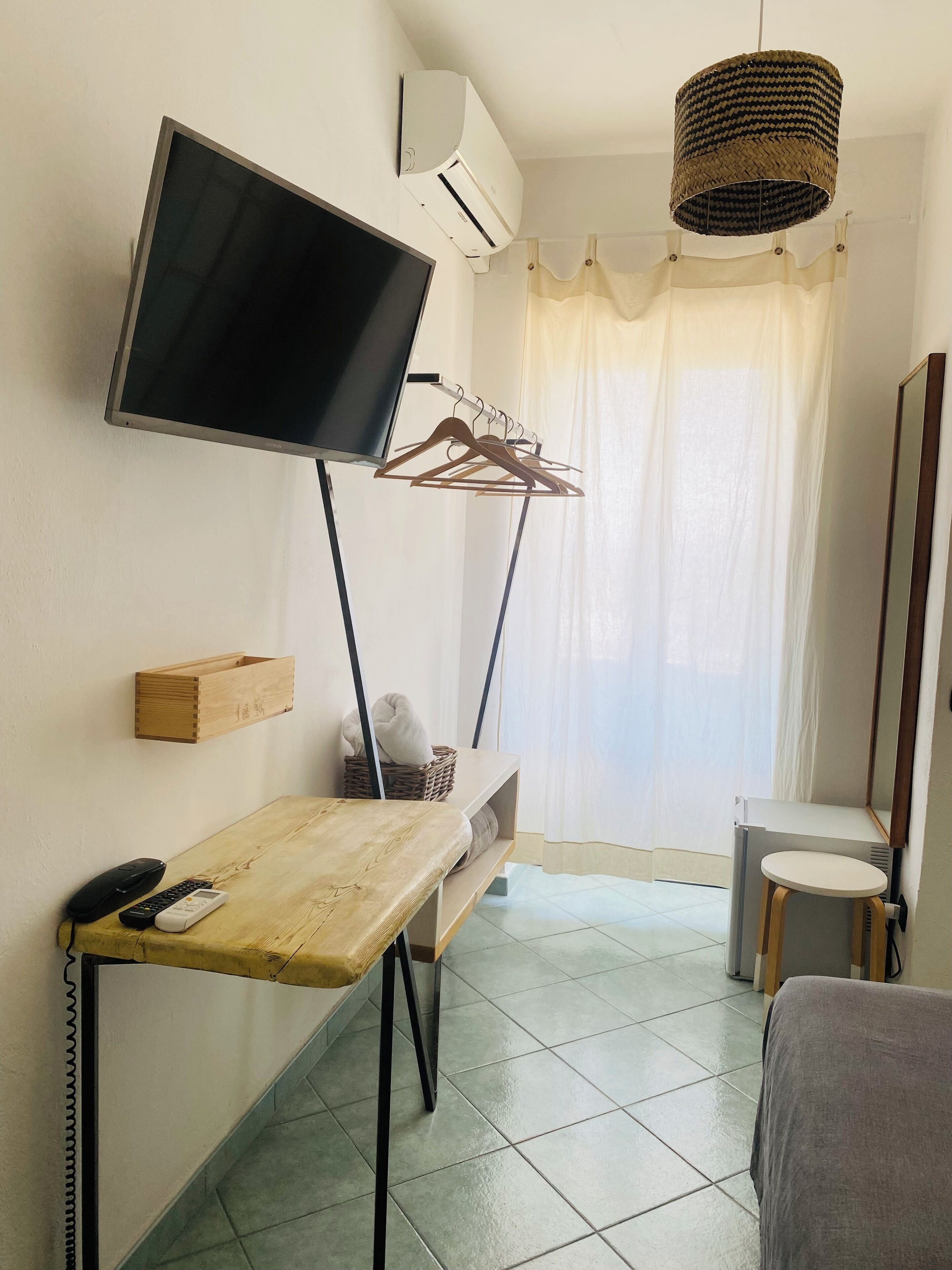 Une chambre d’hôtel compacte, dotée d’un téléviseur à écran plat fixé au mur, d’une table en bois avec une télécommande, d’un petit tabouret et d’un miroir.