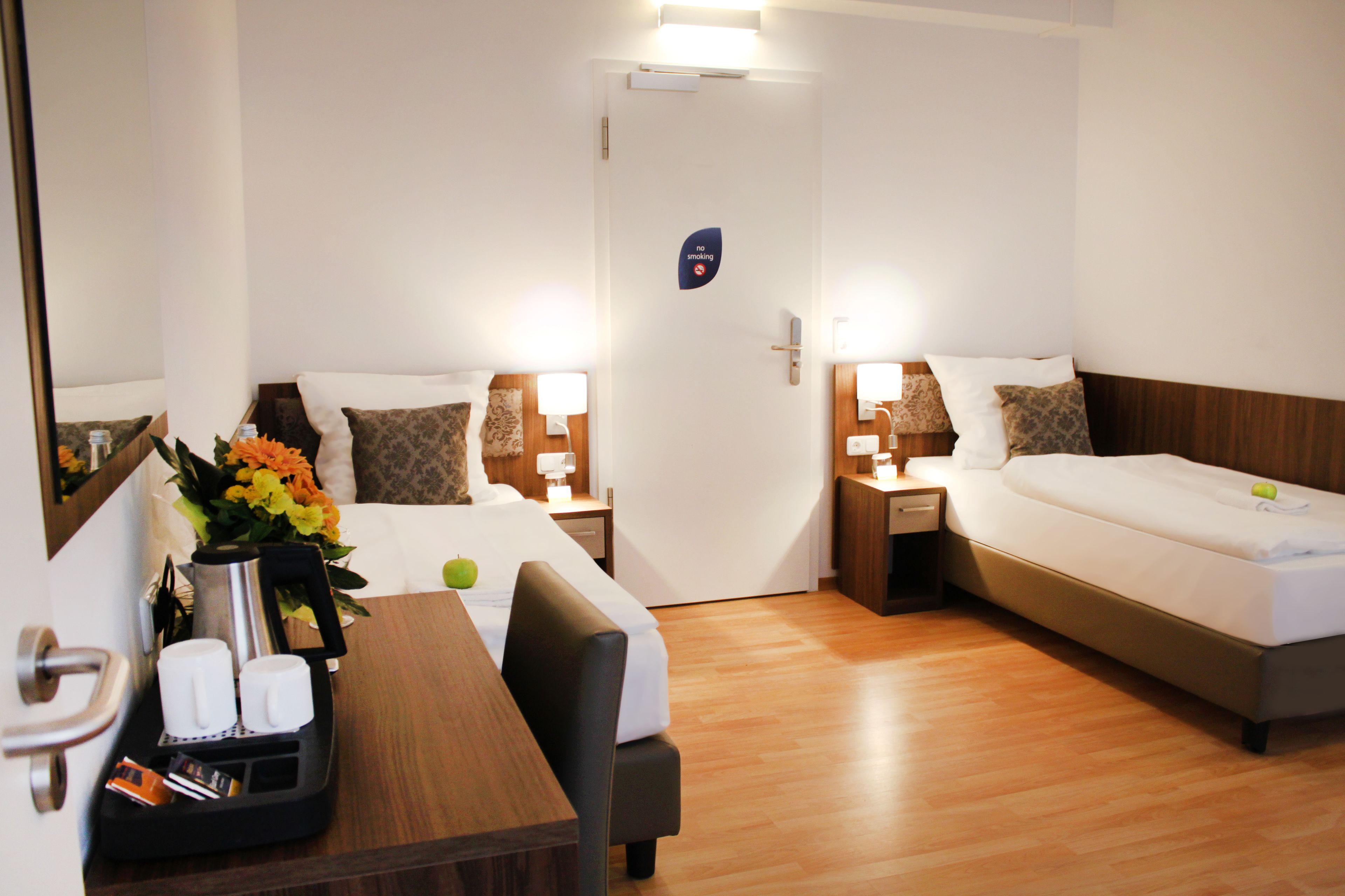 A hotel room with two beds, a desk with a coffee maker, and a "no smoking" sign on the door.
