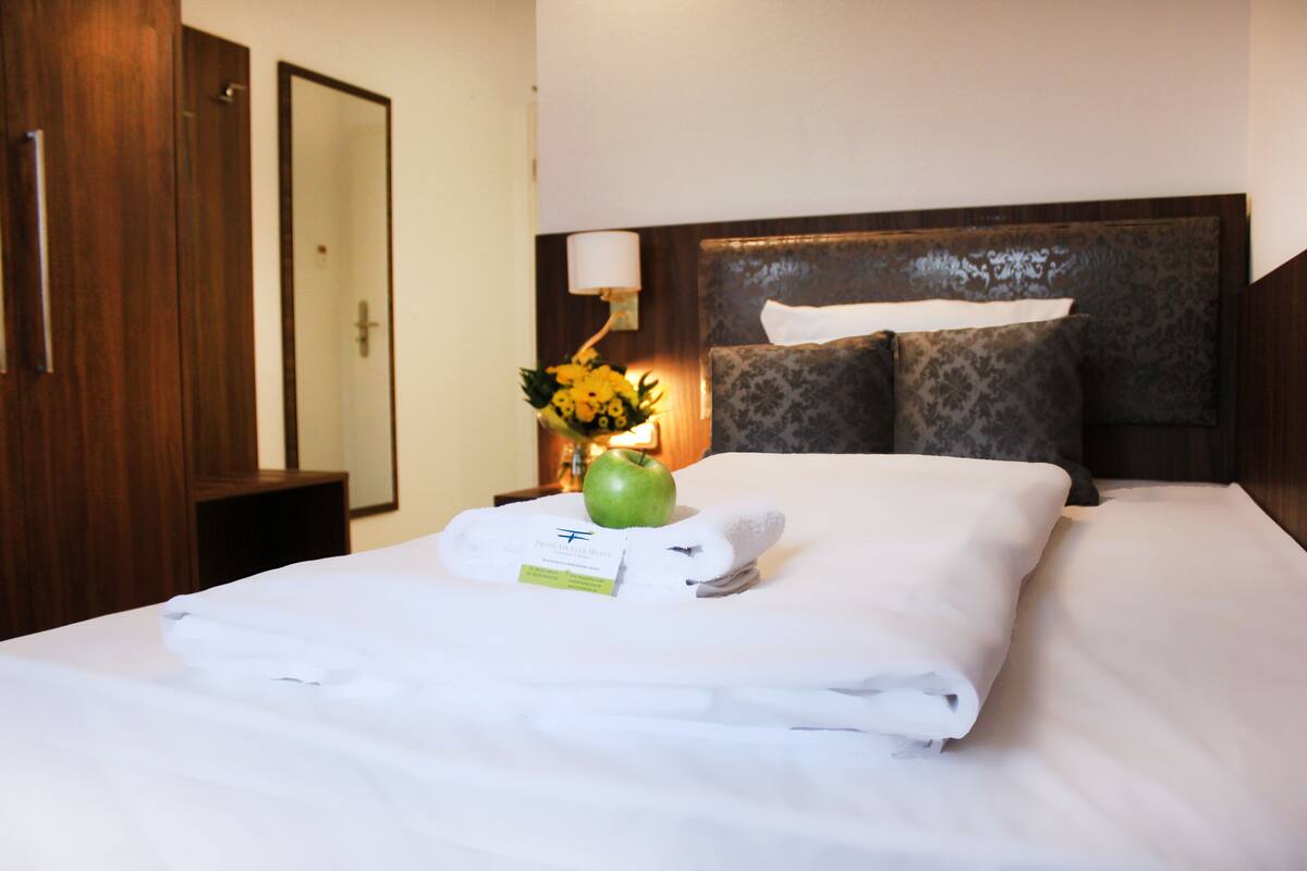 A neatly made bed with a folded towel, a green apple, and a flower arrangement on the nightstand.