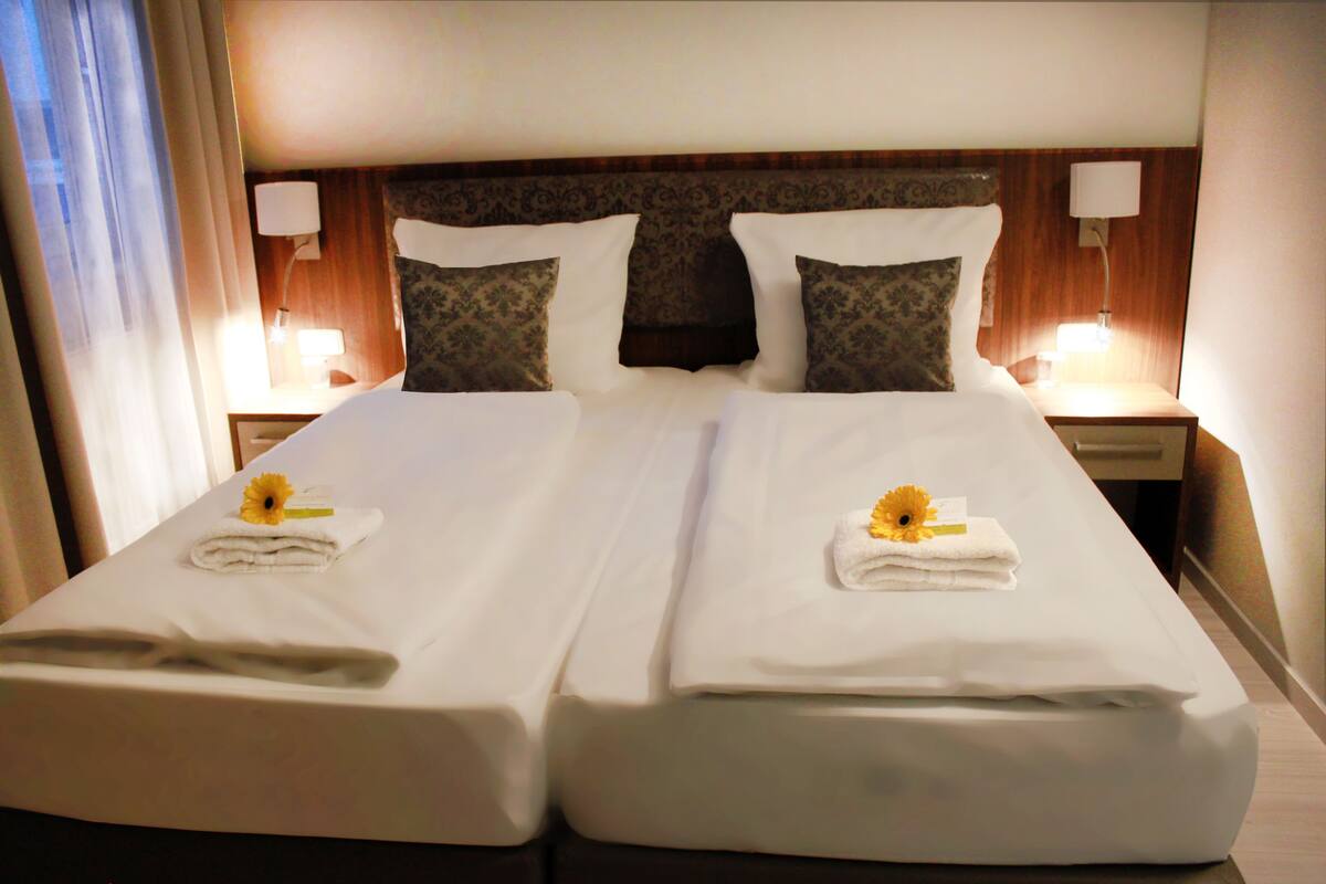 A double bed with white linens, two pillows, and a flower on each pillow. There are two bedside lamps and a wooden headboard.