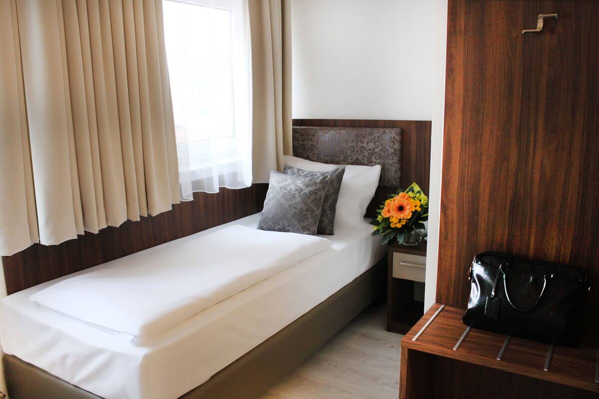 A neatly made bed with white linens, a wooden headboard, and a nightstand with a black handbag and a vase of flowers.