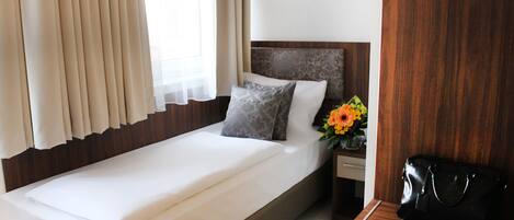 Una cama bien tendida con sábanas blancas, cabecera de madera y mesita de noche con un bolso negro y un florero.