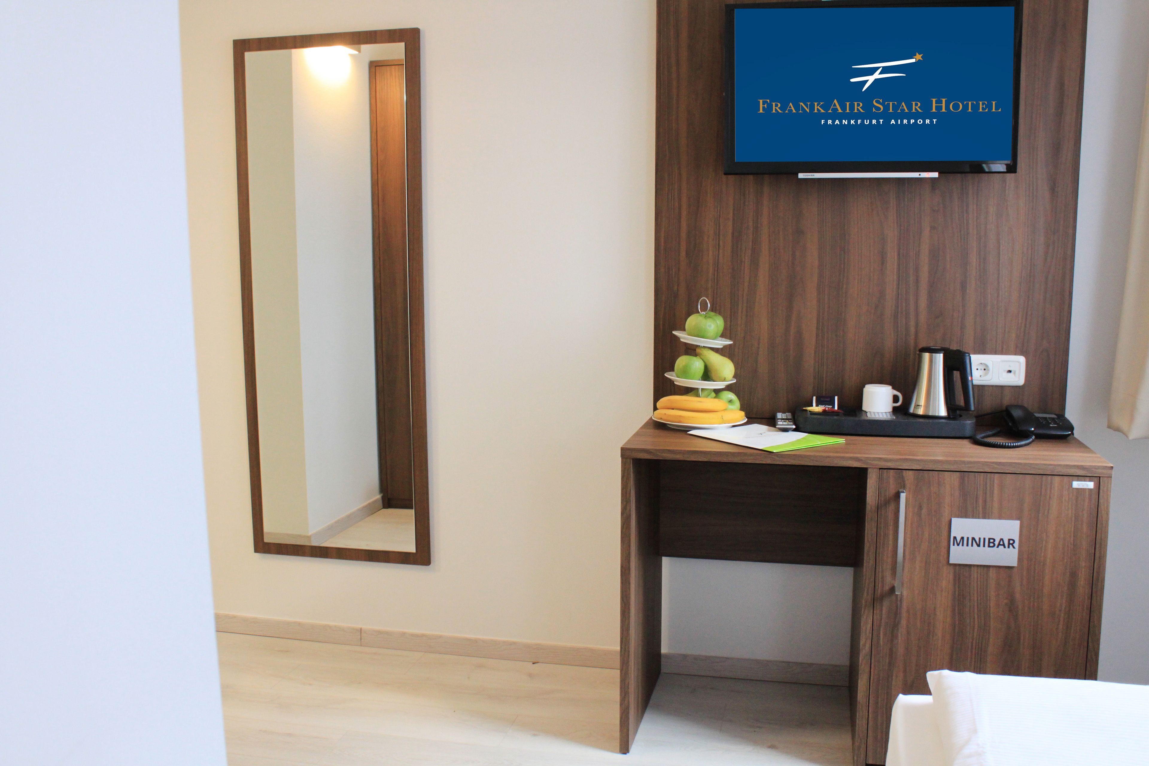 A hotel room with a wooden desk, a minibar, a television displaying the FrankAir Star Hotel logo, and a mirror.