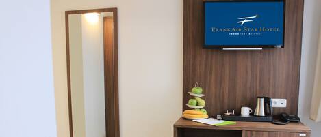 A hotel room with a wooden desk, a minibar, a television displaying the FrankAir Star Hotel logo, and a mirror.