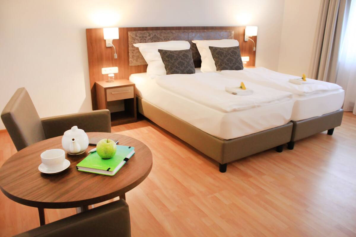 A hotel room with a bed, bedside tables, a chair, a table with a teapot, a cup, and a notebook.