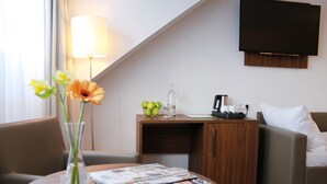 A hotel room with a wooden desk, a sofa, a TV mounted on the wall, and a vase with flowers on the table. - Anor Hotel I Park Sleep & Fly I Frankfurt Airport (Moerfelden-Walldorf)