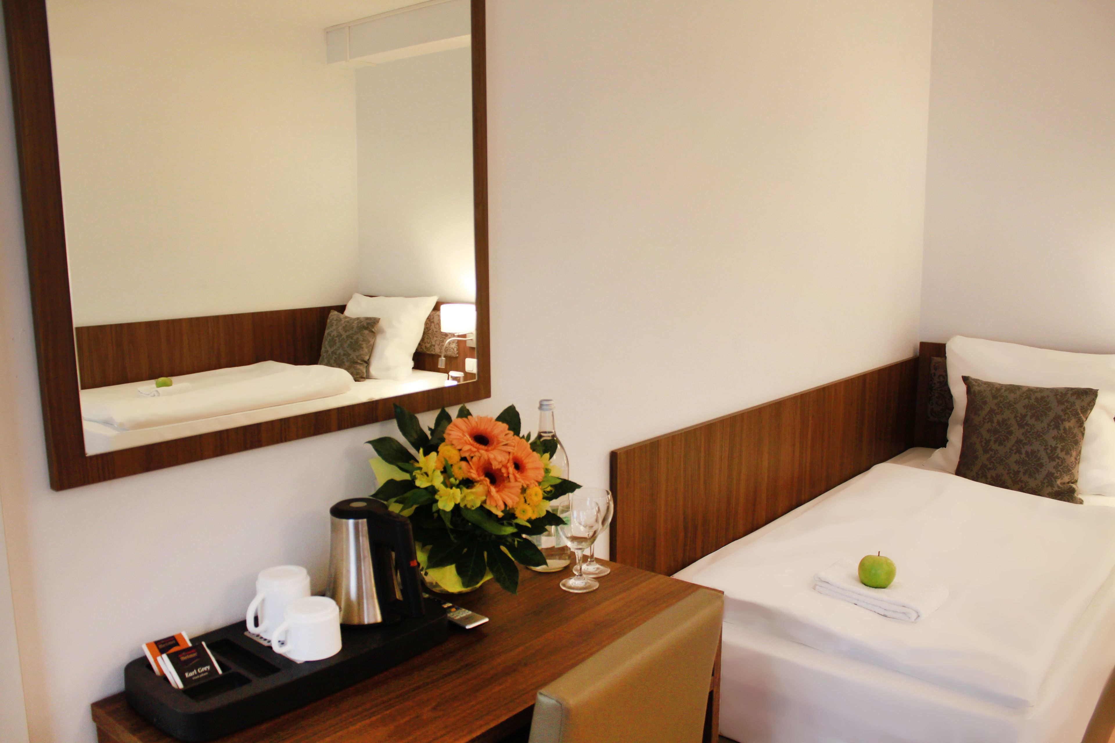 A hotel room with a wooden headboard, a bed with white linens, a bedside table with a flower vase, a coffee maker, and a tray with a glass of water and a remote control.