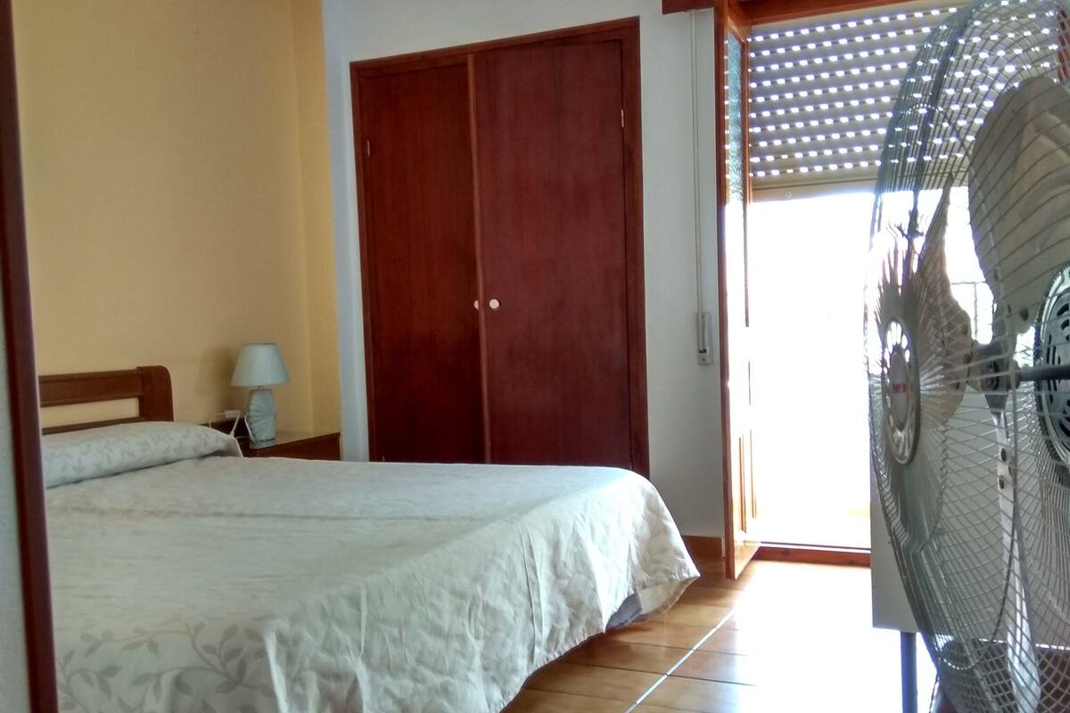 A bedroom with a bed, a nightstand, a lamp, a wooden wardrobe, and a standing fan.
