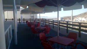 A terrace with red plastic tables and chairs, umbrellas, and a view of the city and hills. - Hospederia Montes (Nijar)