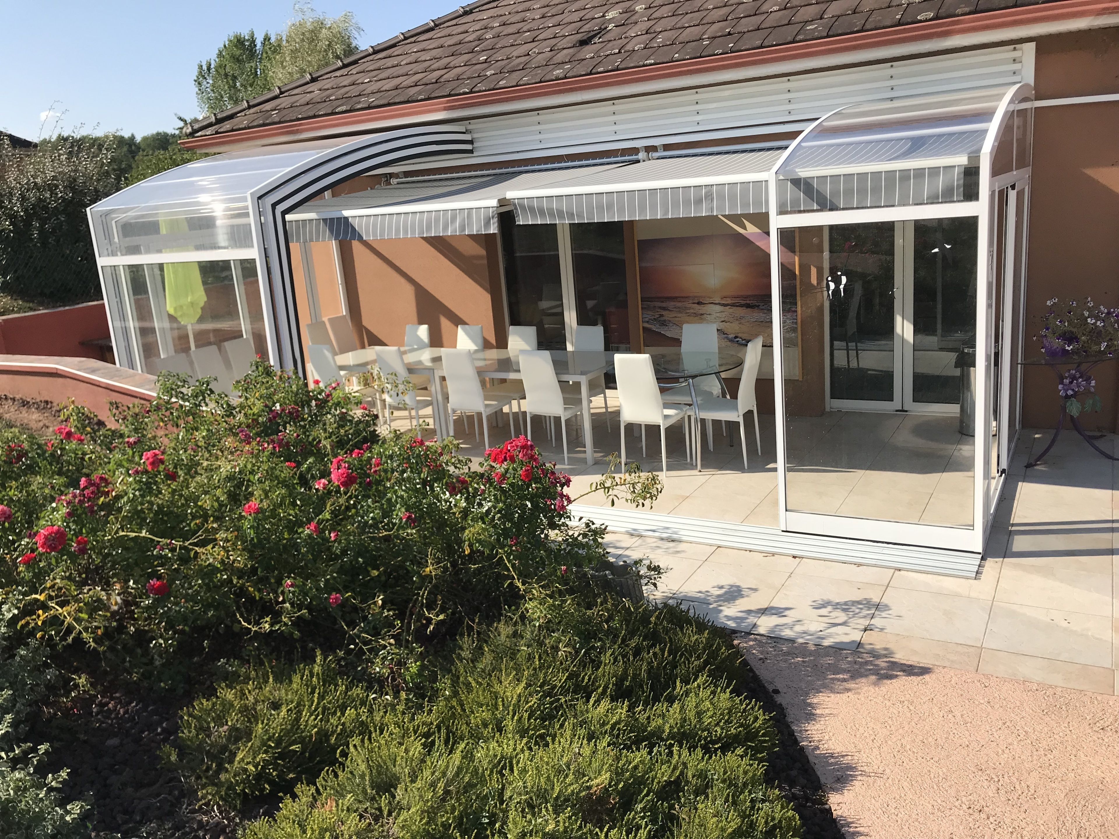 Un espacio comedor al aire libre con paredes de vidrio, sillas blancas y un jardín con flores rosas.