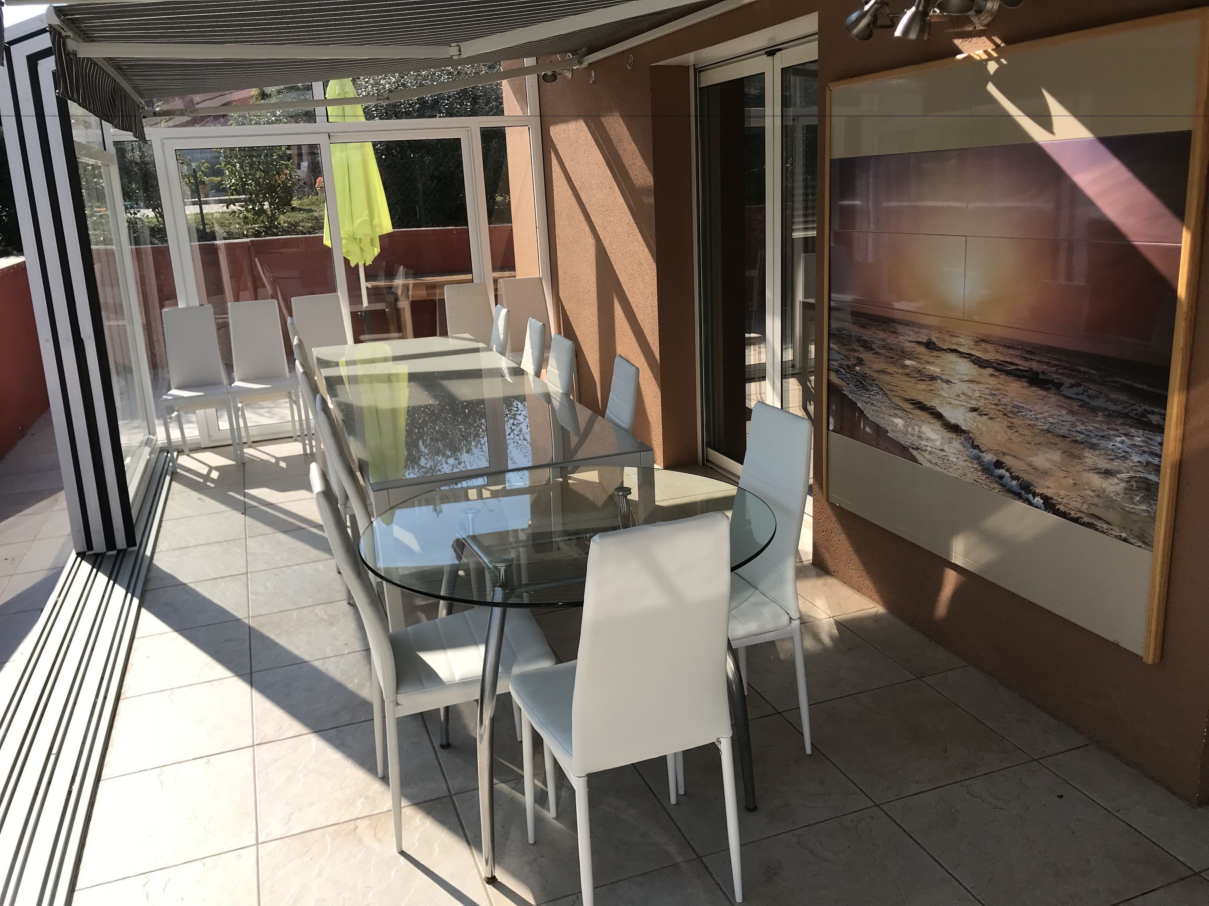 Ein überdachter Patio mit Glaswänden, einem Tisch, Stühlen und einem gelben Sonnenschirm, sowie einem großen Gemälde an der Wand.