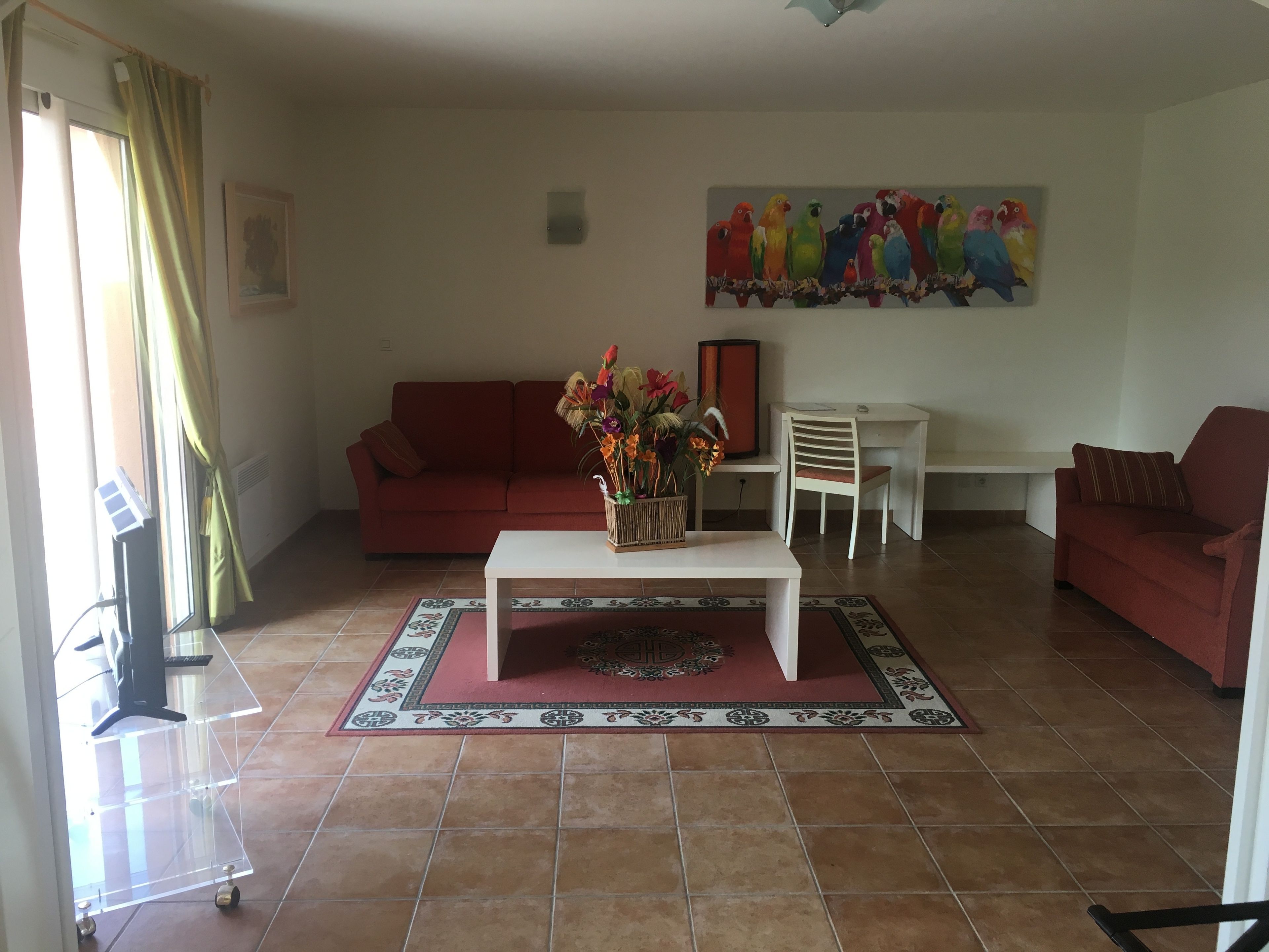 Una sala de estar con un sofá rojo, una mesa de centro blanca con un florero, un escritorio blanco con una silla, y un cuadro de un loro colorido en la pared.