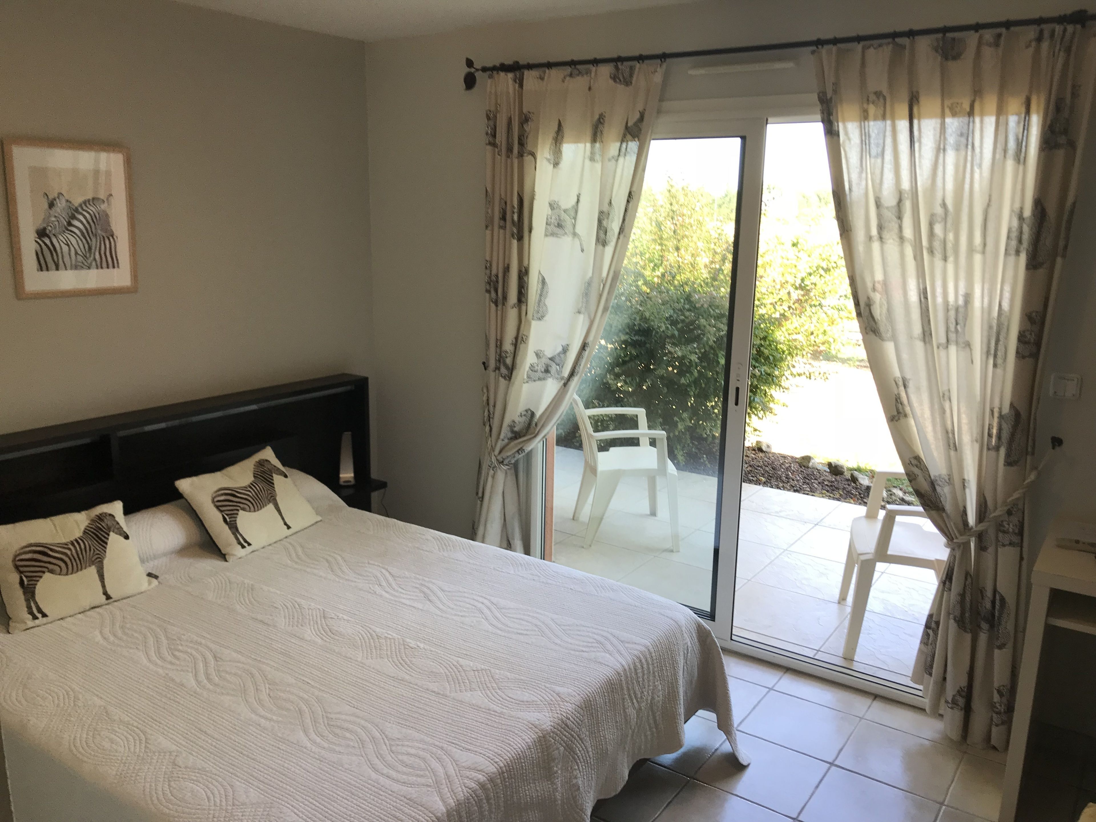 Une chambre à coucher avec un lit, des coussins à motifs zébrés, un tableau encadré accroché au mur et une porte-fenêtre coulissante donnant sur un espace extérieur équipé d’une chaise.