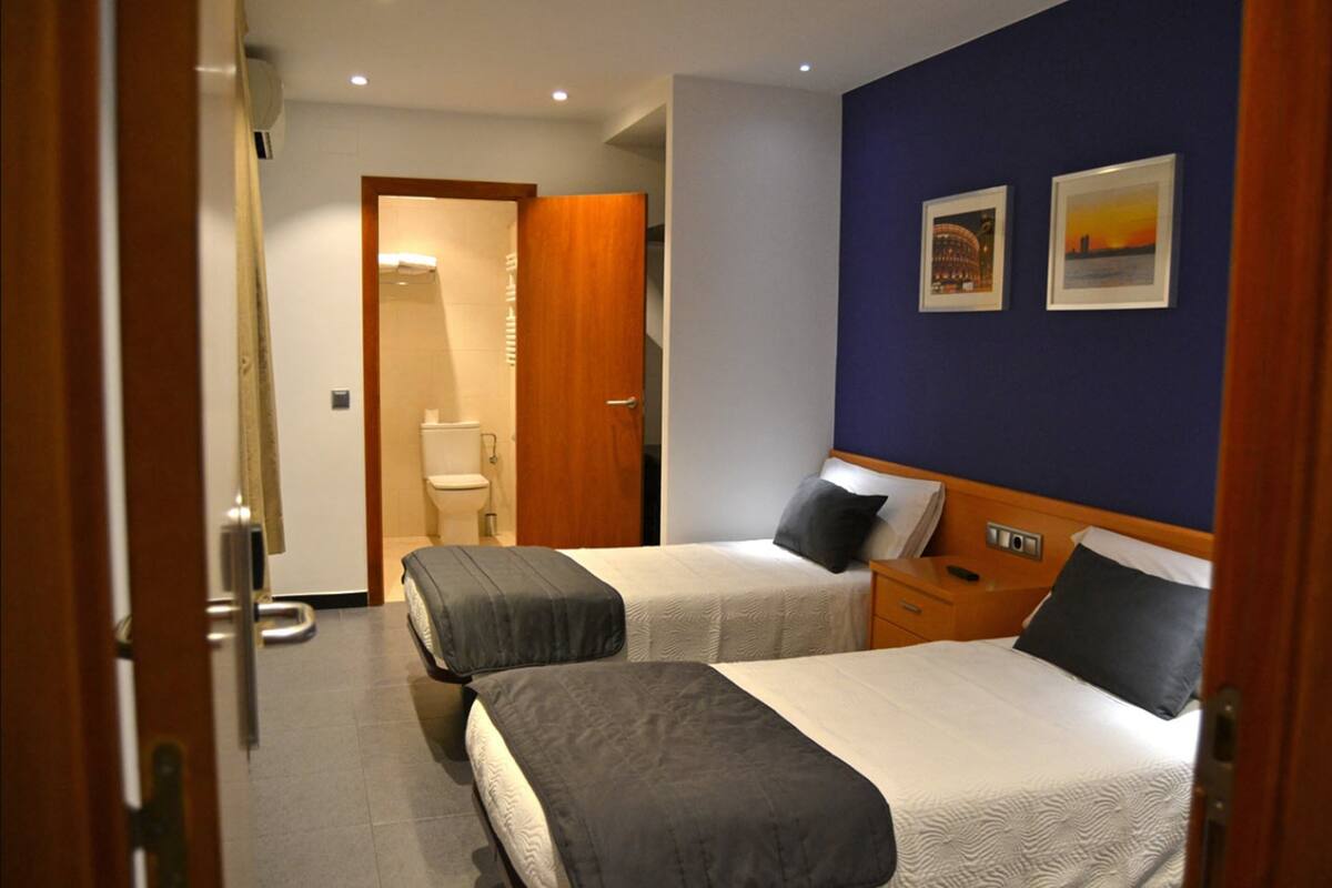 Habitación de hotel con dos camas, una mesita de noche de madera, un baño con inodoro y dos cuadros enmarcados en la pared.