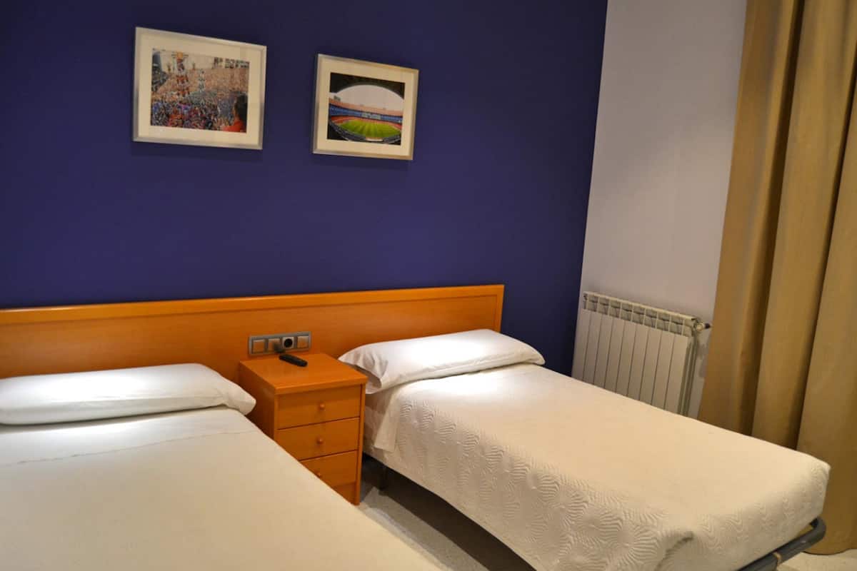 Habitación de hotel con dos camas individuales, cabecero de madera, mesita de noche con un control remoto y dos cuadros en la pared.