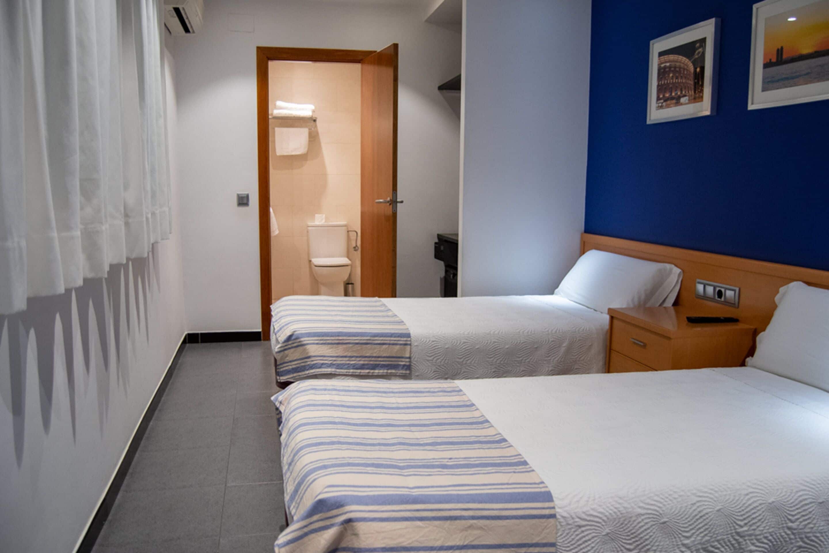 A hotel room with two beds, a wooden nightstand, a bathroom with a toilet, and a door leading to another room.