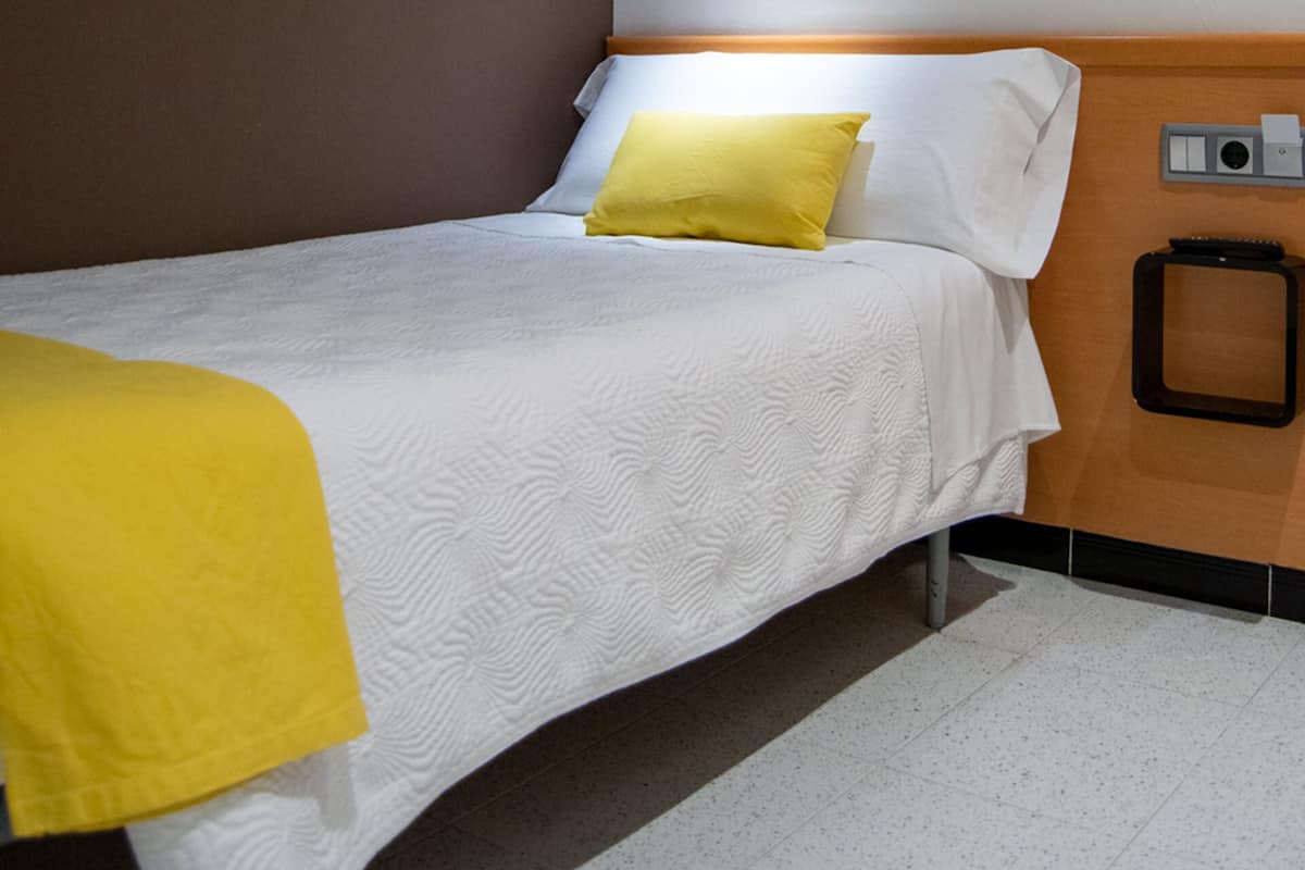 Una cama individual con ropa de cama blanca y una manta amarilla, una mesita de noche con lámpara, y un despertador fijado a la pared.
