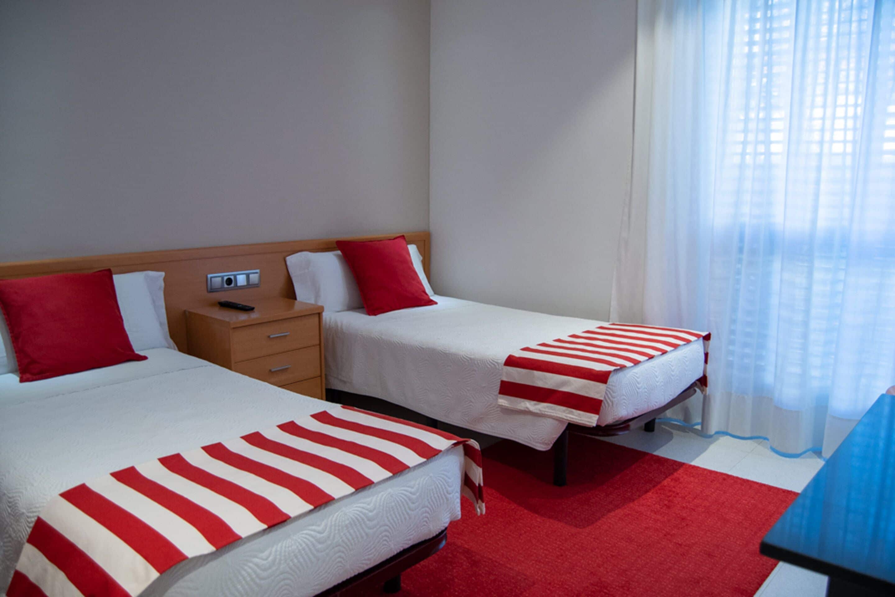 Habitación de hotel con dos camas, cada una con ropa de cama a rayas rojas y blancas, una mesita de noche de madera y una alfombra roja.