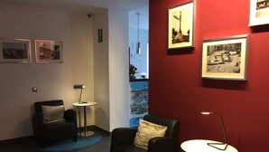 A room with a red accent wall, a black leather chair, a small round table, and a blue rug. There are framed pictures on the walls and a window with a view of a building. - Hostal Apolo (Barcelona)