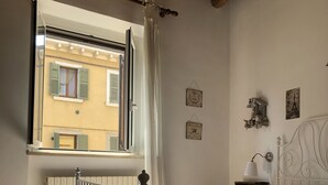 A bedroom with a bed, a window with curtains, a radiator, a bedside table with a lamp, and wall decorations. - Ai Leoni (Verona)